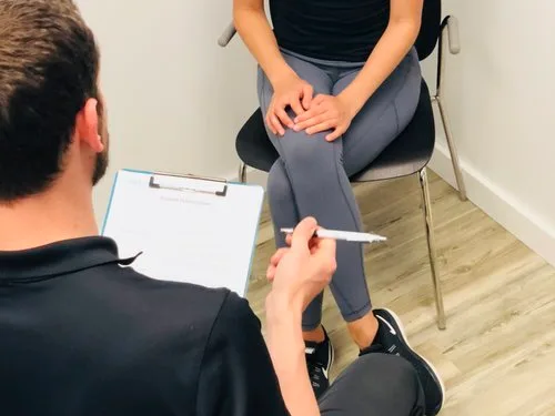 A Radius provider holds a clipboard and pen during an intake consultation with a seated patient in athletic wear