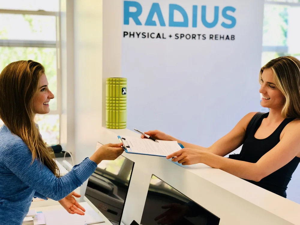 A smiling Radius front-desk team member hands a patient an intake clipboard across the reception counter, beneath the Radius Physical + Sports Rehab logo