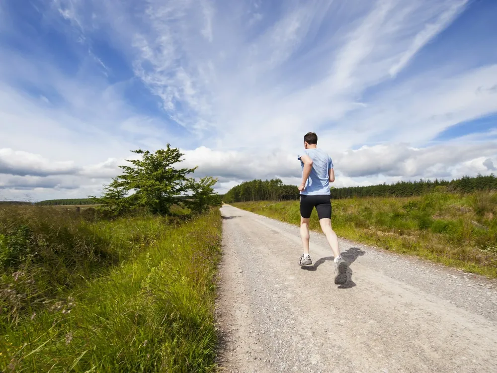 A runner seen from behind jogging along a gravel trail through open grassy countryside under a bright blue, cloud-filled sky